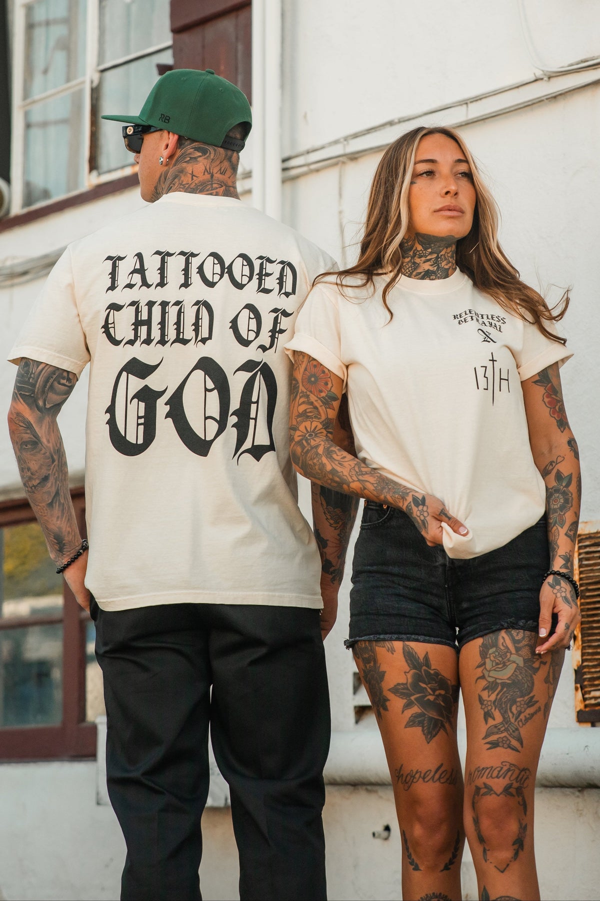 Two tattooed individuals in casual outfits stand outdoors; the man wears the Relentless Betrayal Tattooed Child of God Premium Ivory T-Shirt—a striking piece of Christian apparel—while the woman sports a shirt that says Wolvesville 13th.