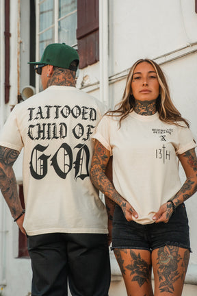 Two tattooed models pose in casual outfits; the woman stands facing forward in black shorts, while the man turns his back to showcase the “Tattooed Child of God Premium Ivory T-Shirt” by Relentless Betrayal—a bold statement piece from our Christian apparel collection.