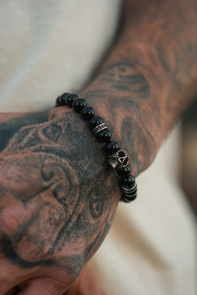 A tattooed hand adorned with the Relentless Betrayal Day of the Dead Black Bead Bracelet, featuring metallic accents and a small skull charm that captures the festive spirit of Día de los Muertos.