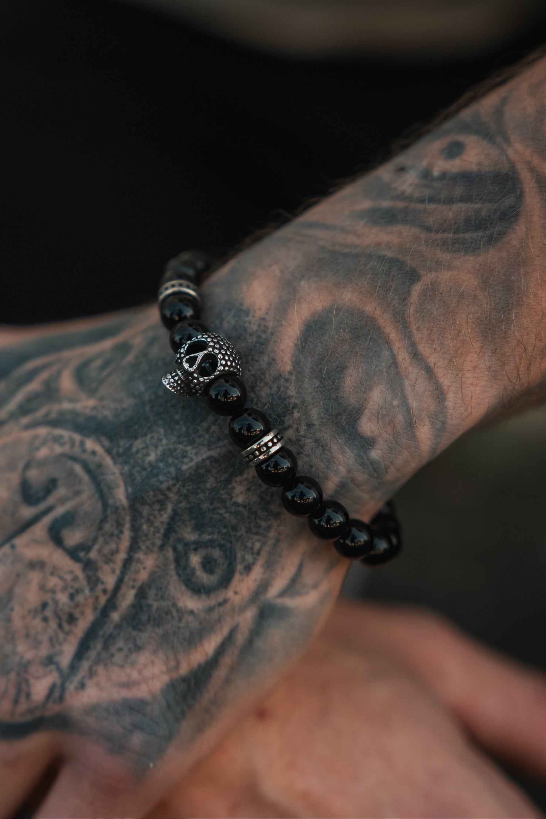 A tattooed arm displays the Relentless Betrayal Day of the Dead Black Bead Bracelet in close-up detail, showcasing its striking silver skull accent and intricate semi-precious gemstone design.
