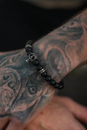 A tattooed arm displays the Relentless Betrayal Day of the Dead Black Bead Bracelet in close-up detail, showcasing its striking silver skull accent and intricate semi-precious gemstone design.