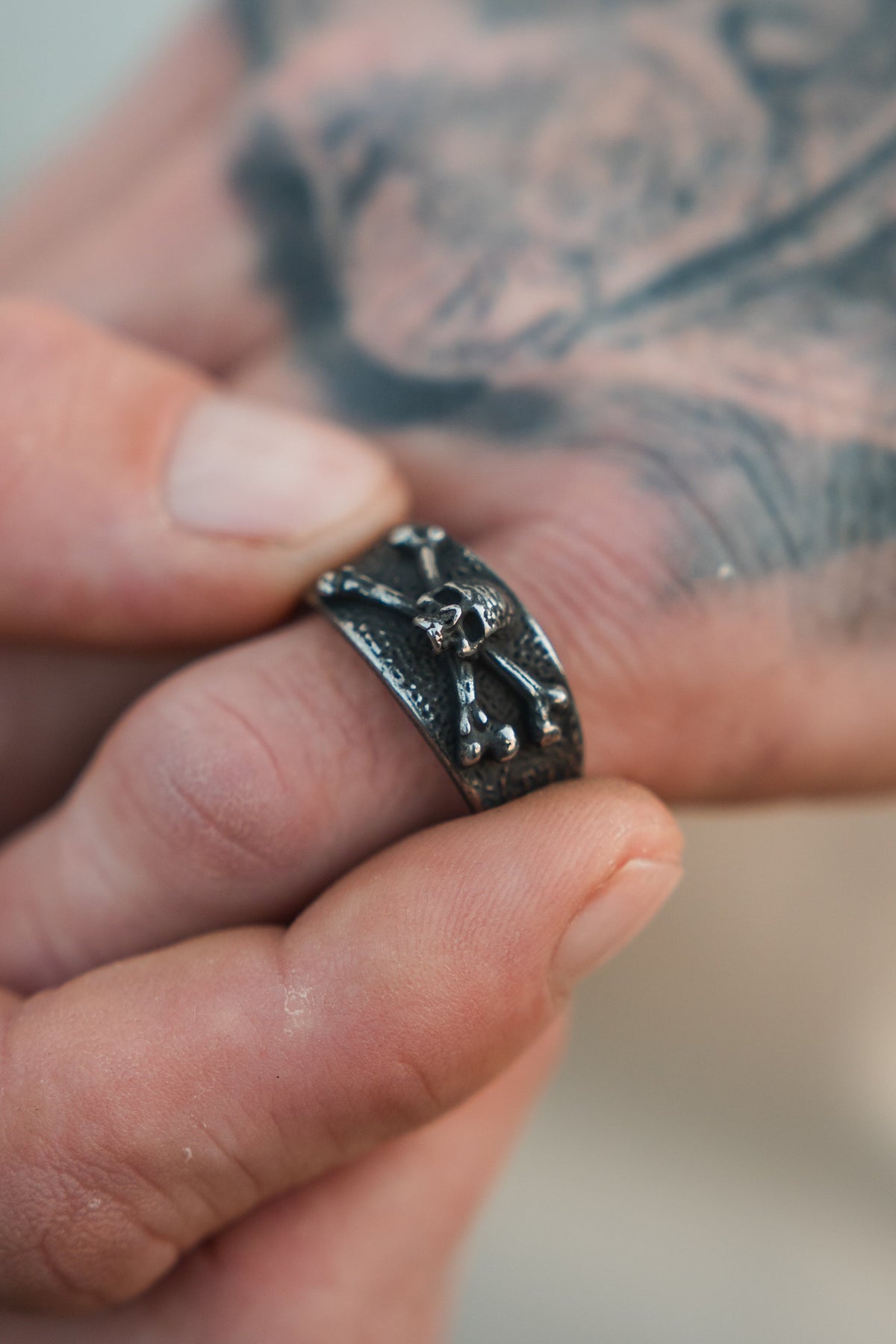 A tattooed hand holds the Relentless Betrayal Bone Crusher Ring, a stainless steel piece adorned with raised animal and bone details.