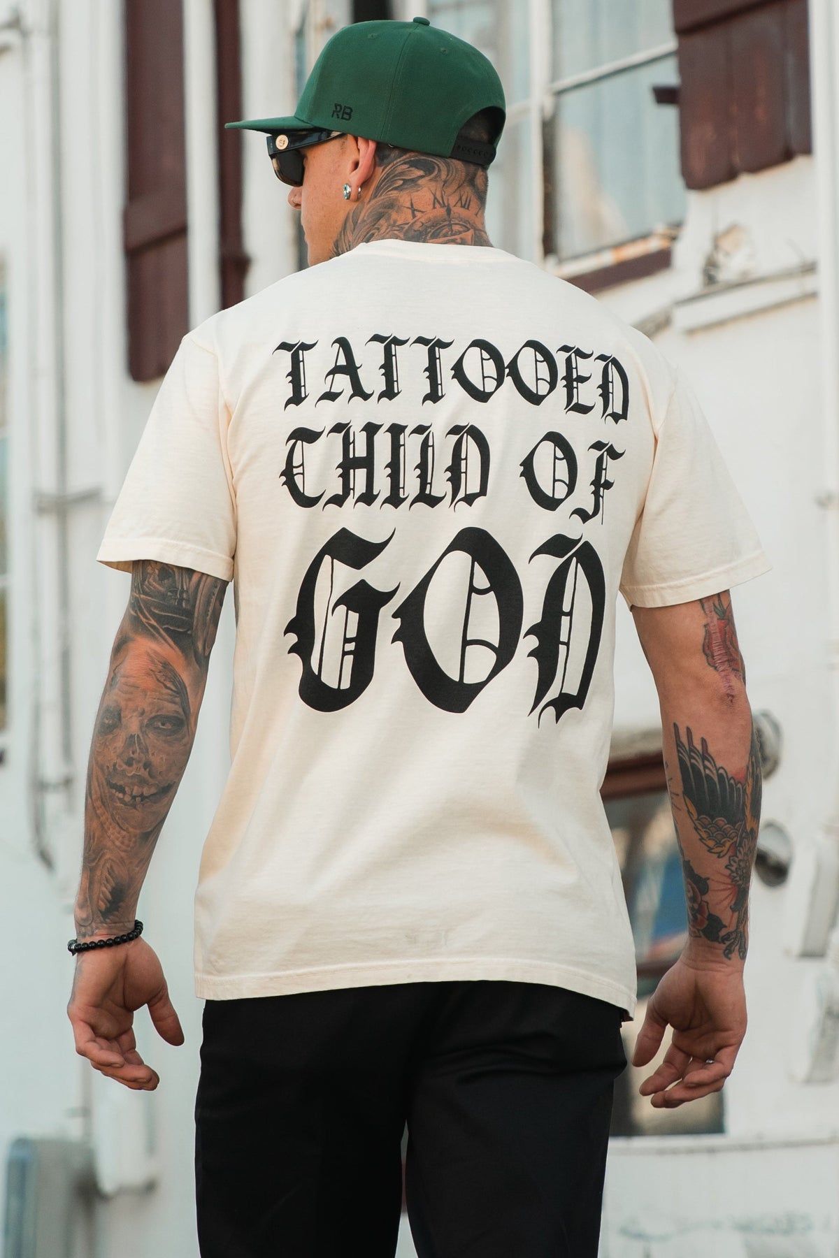 A tattooed individual wearing the Relentless Betrayal Tattooed Child of God Premium Ivory T-Shirt, featuring bold black lettering on the back, stands outdoors and proudly displays striking Christian apparel.