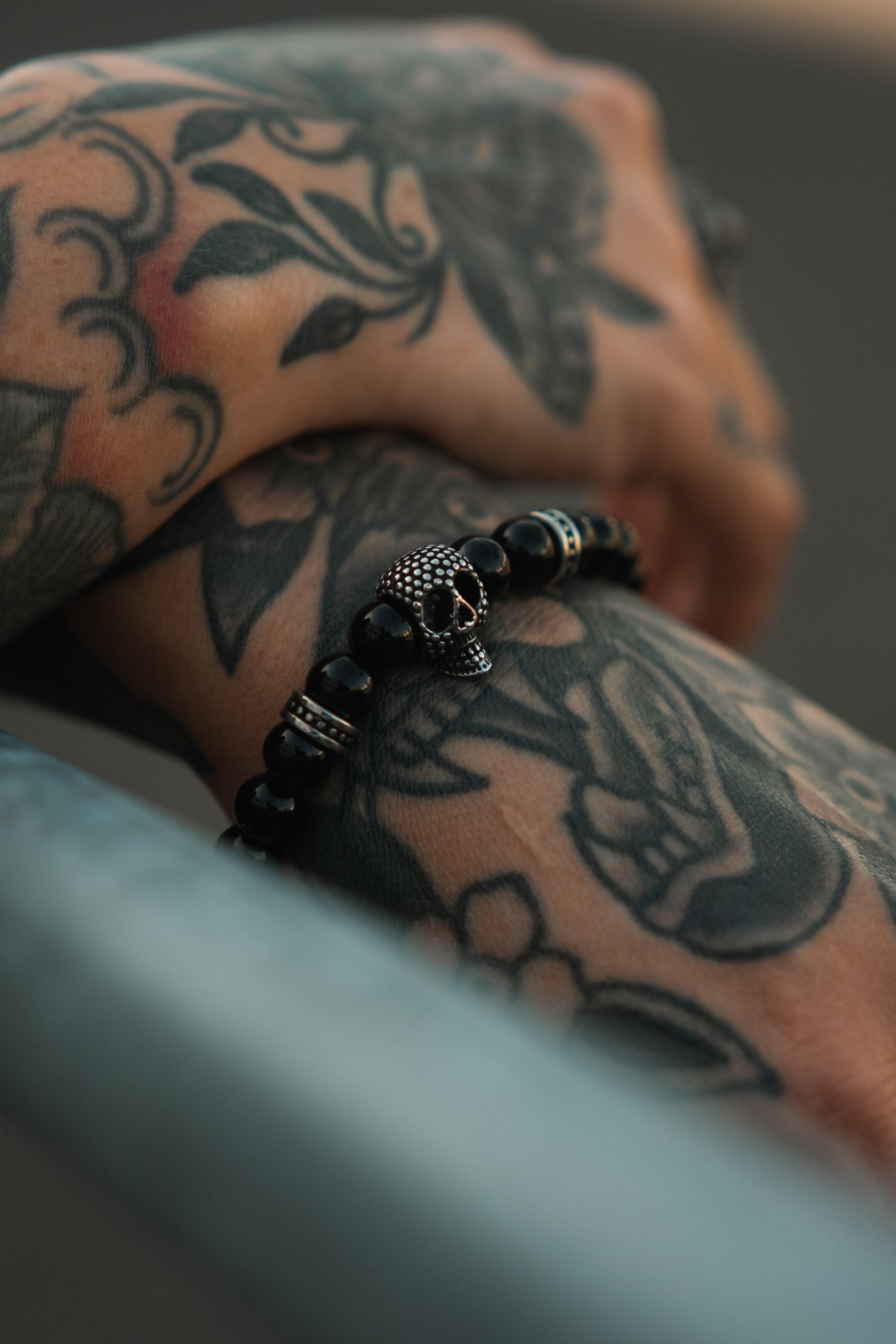 Tattooed hands rest on a metal surface, adorned with the Relentless Betrayal Day of the Dead Black Bead Bracelet—its striking black beads and skull charm capturing bold, artistic style.