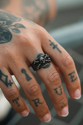 A tattooed hand with "TRUE" on the fingers wears the Relentless Betrayal Bone Crusher Ring, a dark metal pirate-inspired piece, along with another textured stainless steel ring. More tattoos cover the knuckles and back of the hand.