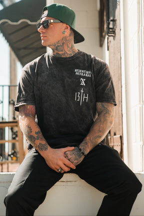 A tattooed man in a Tattooed Child of God Premium Mineral Wash T-Shirt by Relentless Betrayal, black pants, green cap, and sunglasses sits outdoors with clasped hands, looking to the side. The shirt features a small graphic and “13TH.”.