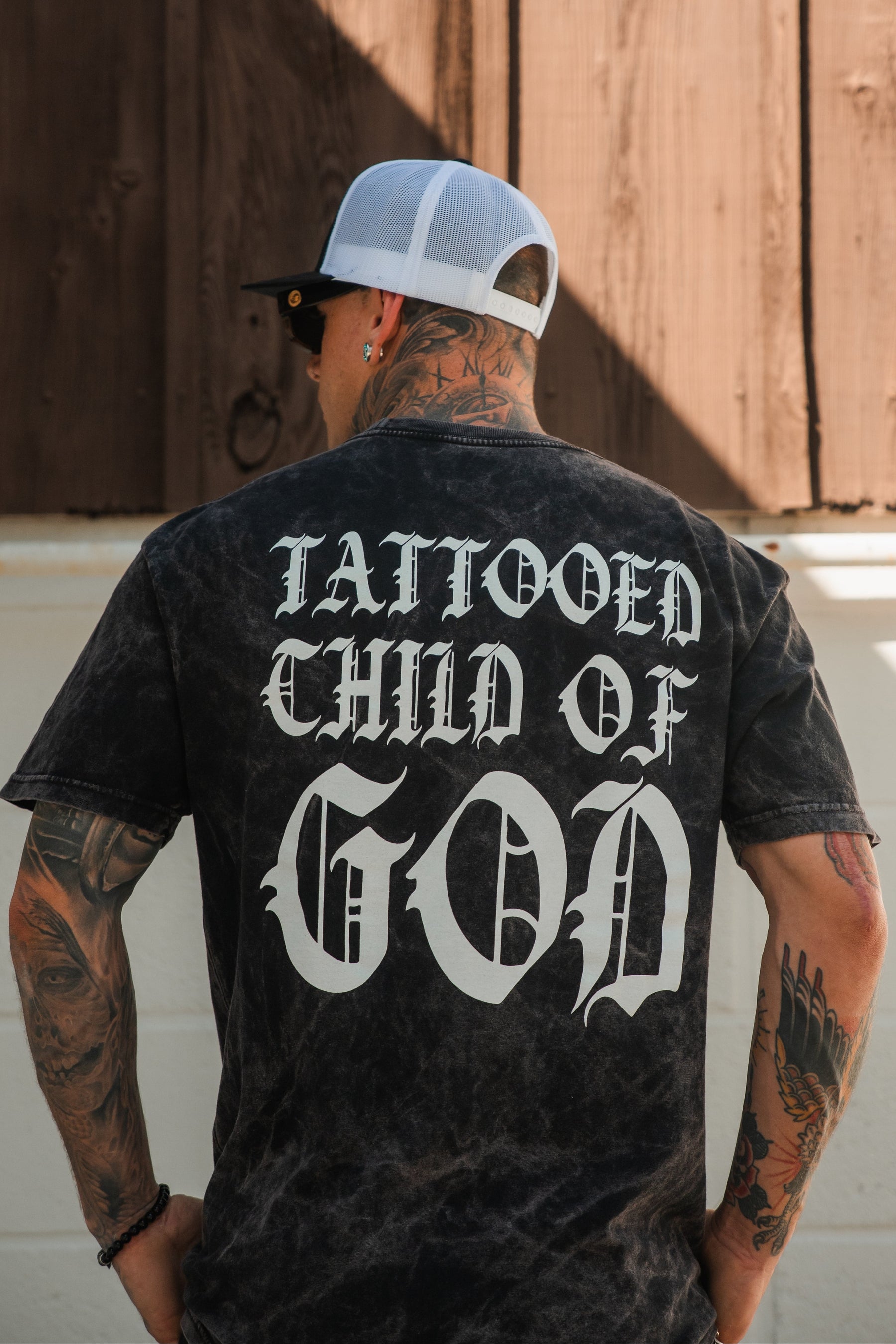 A tattooed person models the Relentless Betrayal Tattooed Child of God Premium Mineral Wash T-Shirt, featuring bold white gothic text on the back, while standing outdoors and showcasing distinctive Christian style.