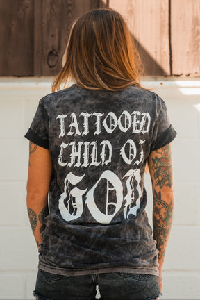 Seen from behind, a person with tattooed arms wears the Relentless Betrayal Tattooed Child of God Premium Mineral Wash T-Shirt, featuring bold white text and standing out as unique Christian apparel.