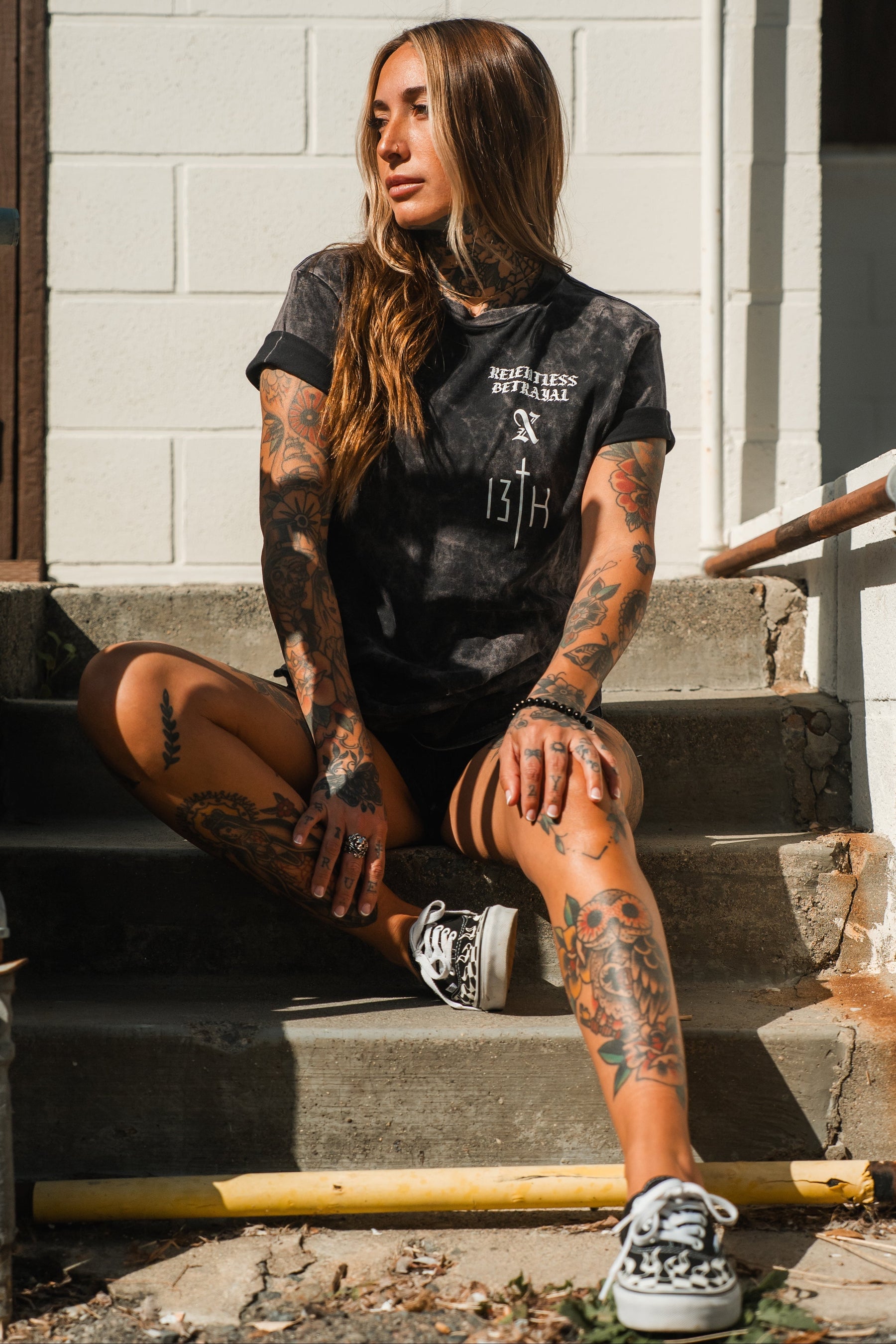 A woman with long hair and extensive tattoos sits on outdoor steps, wearing the Relentless Betrayal Tattooed Child of God Premium Mineral Wash T-Shirt, black shorts, and patterned sneakers as she looks to the side in bright sunlight.