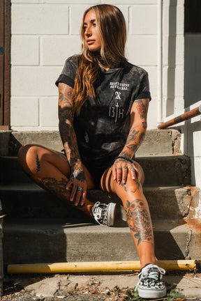 A woman with long hair and extensive tattoos sits on outdoor steps, wearing the Relentless Betrayal Tattooed Child of God Premium Mineral Wash T-Shirt, black shorts, and patterned sneakers as she looks to the side in bright sunlight.
