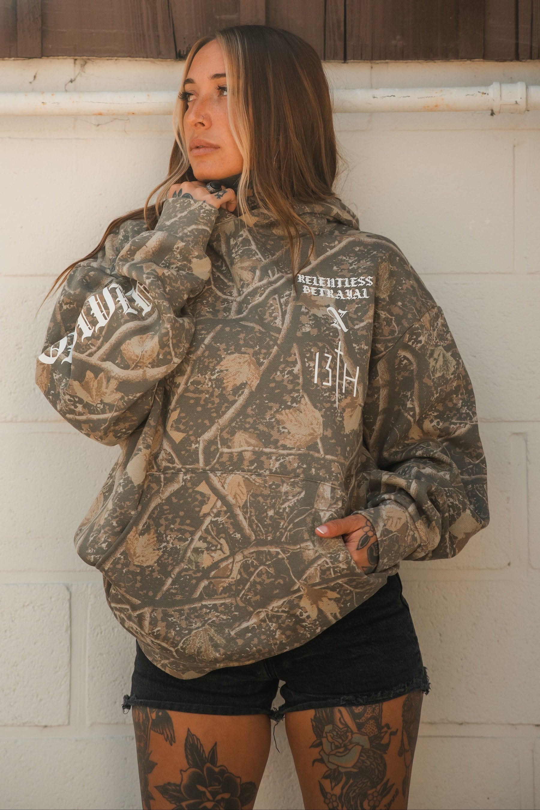 Person wearing a Tattooed Child of God Premium Deadwood Camo Hoodie with white text and graphics on the chest and sleeve.
