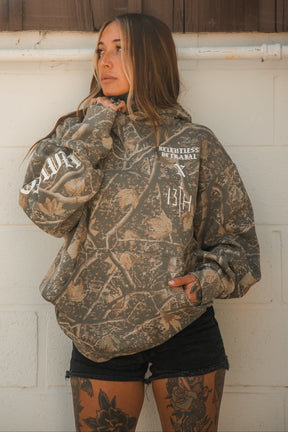 Person wearing a Tattooed Child of God Premium Deadwood Camo Hoodie with white text and graphics on the chest and sleeve.