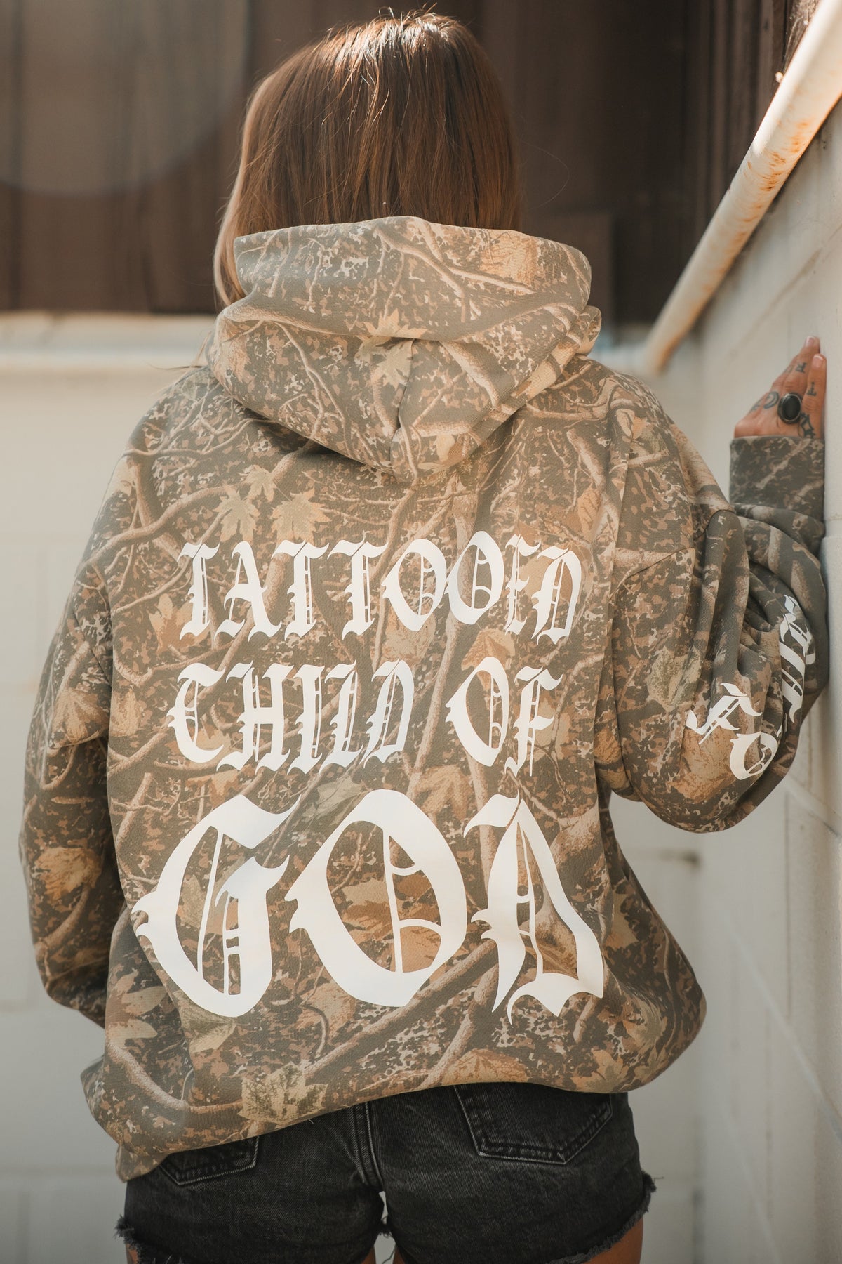 Person wearing a Tattooed Child of God Premium Deadwood Camo Hoodie with the text printed in white gothic lettering on the back.
