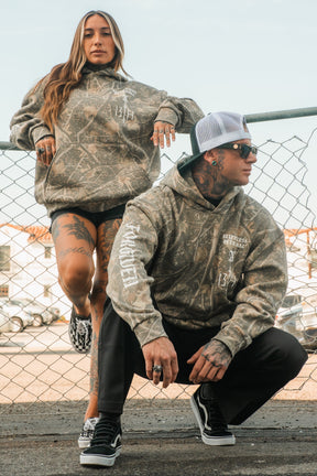 Two people wearing matching Tattooed Child of God Premium Deadwood Camo Hoodies leaning against a chain-link fence.
