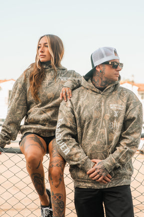 A woman and a man wearing matching Tattooed Child of God Premium Deadwood Camo Hoodies by Relentless Betrayal, leaning against a chain-link fence.