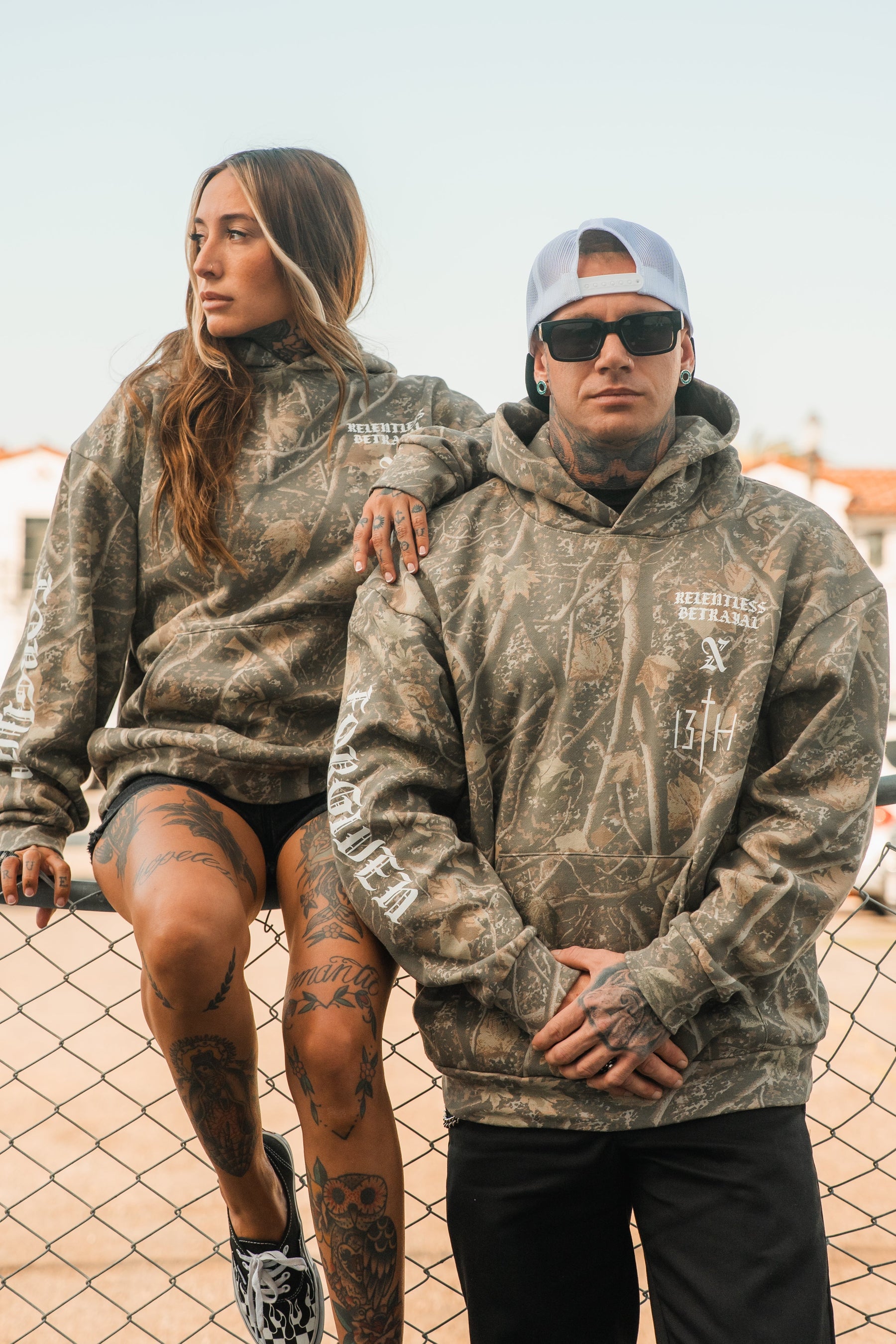 A man and woman pose by a chain-link fence wearing matching Tattooed Child of God Premium Deadwood Camo Hoodies.