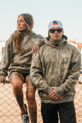 A man and woman pose by a chain-link fence wearing matching Tattooed Child of God Premium Deadwood Camo Hoodies.