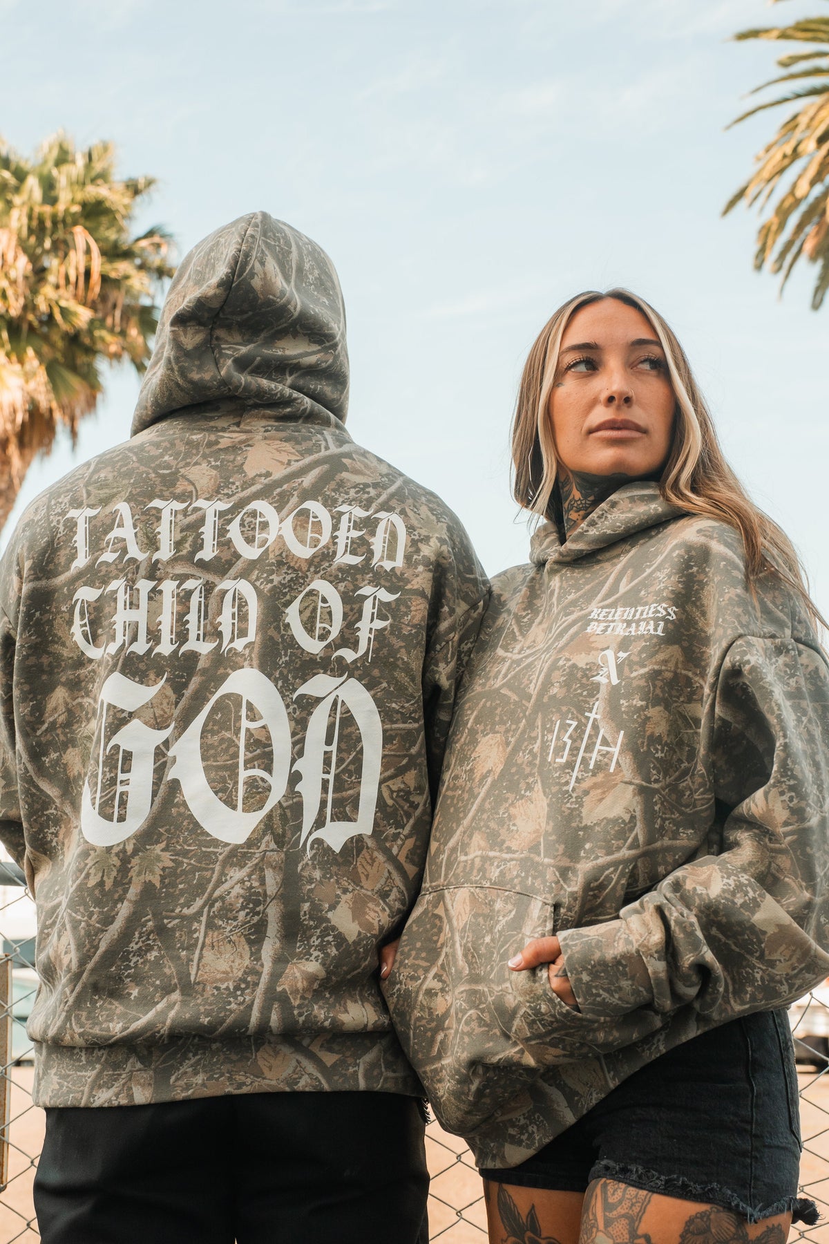 Two people wear matching Tattooed Child of God Premium Deadwood Camo Hoodies; the back of one features the text TATTOOED CHILD OF GOD in large white lettering.