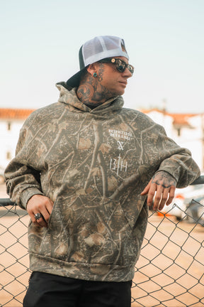 A person in a Tattooed Child of God Premium Deadwood Camo Hoodie and a baseball cap leans against a chain-link fence.