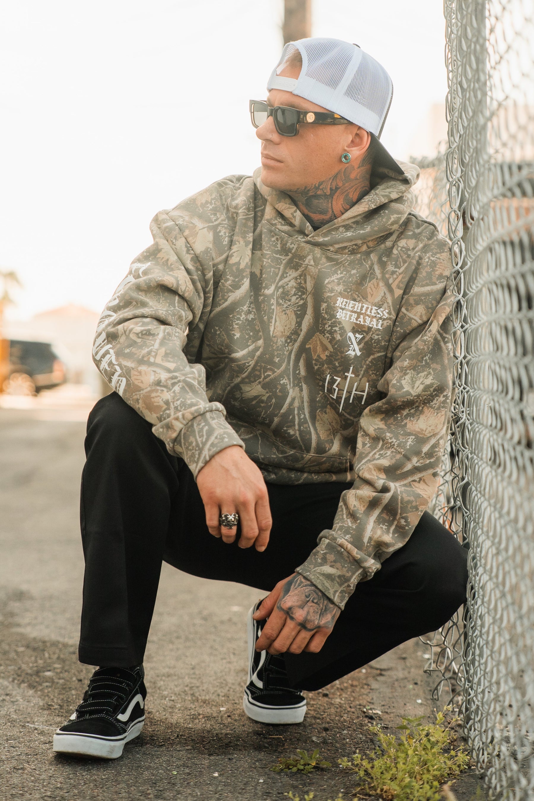 A man in a Tattooed Child of God Premium Deadwood Camo Hoodie, black trousers, black sneakers, a baseball cap, and sunglasses crouches next to a chain-link fence.