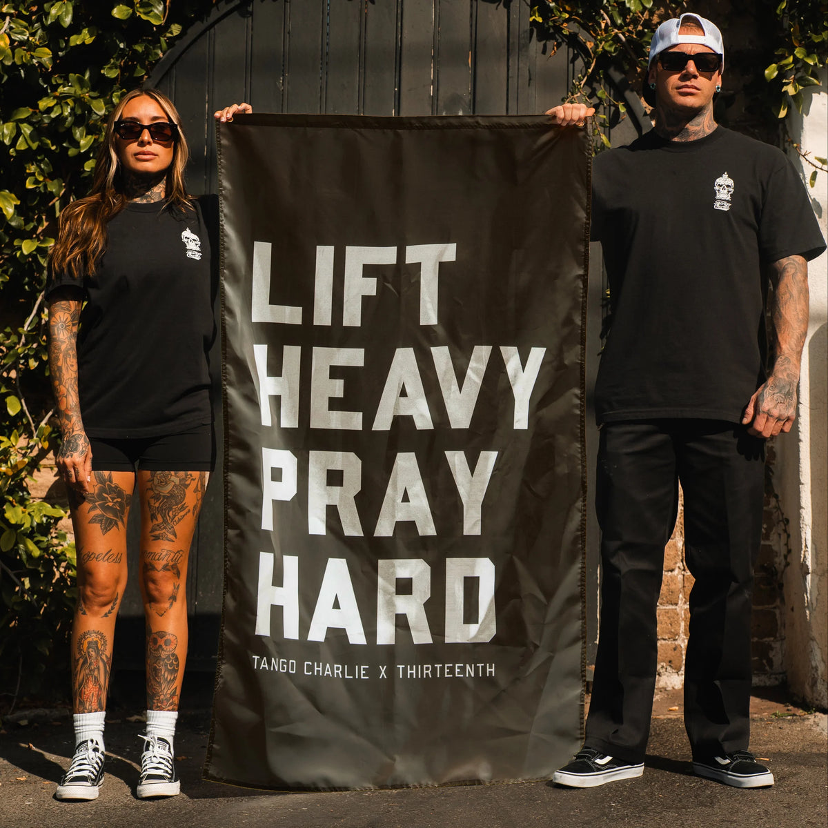 Two people in black outfits and sunglasses stand outdoors, holding the "Lift heavy. Pray hard." Black Flag by Tango Charlie Apparel—the perfect motivational gym banner.