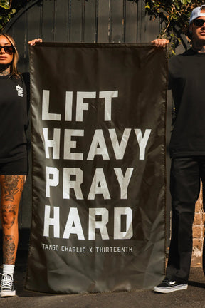 Two people in black outfits and sunglasses stand outdoors, holding the "Lift heavy. Pray hard." Black Flag by Tango Charlie Apparel—the perfect motivational gym banner.