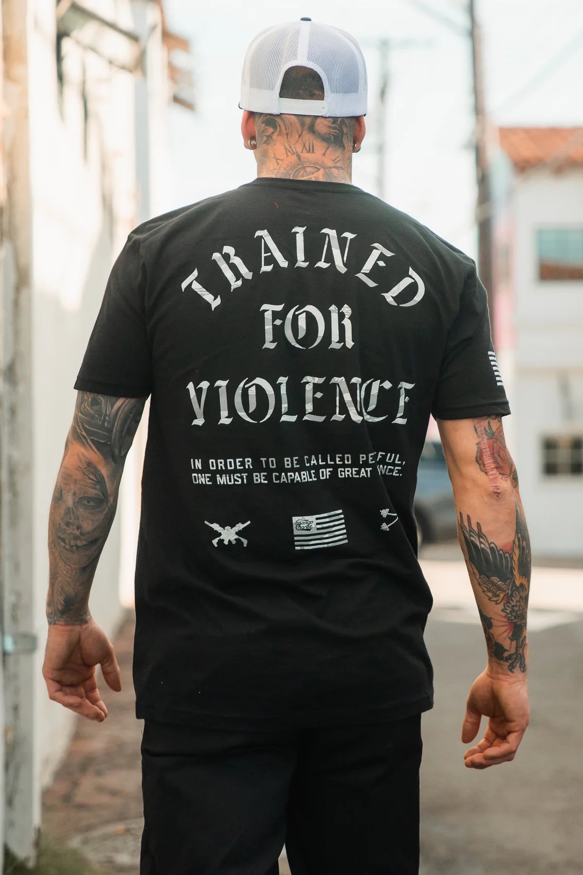 A tattooed man in an alley wears the Tango Charlie Apparel "Trained for Violence - Tee," a black shirt with bold text and small graphics, reflecting psychological conditioning and technical skill.