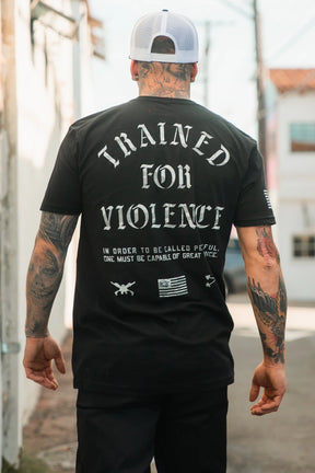 A tattooed man in an alley wears the Tango Charlie Apparel "Trained for Violence - Tee," a black shirt with bold text and small graphics, reflecting psychological conditioning and technical skill.