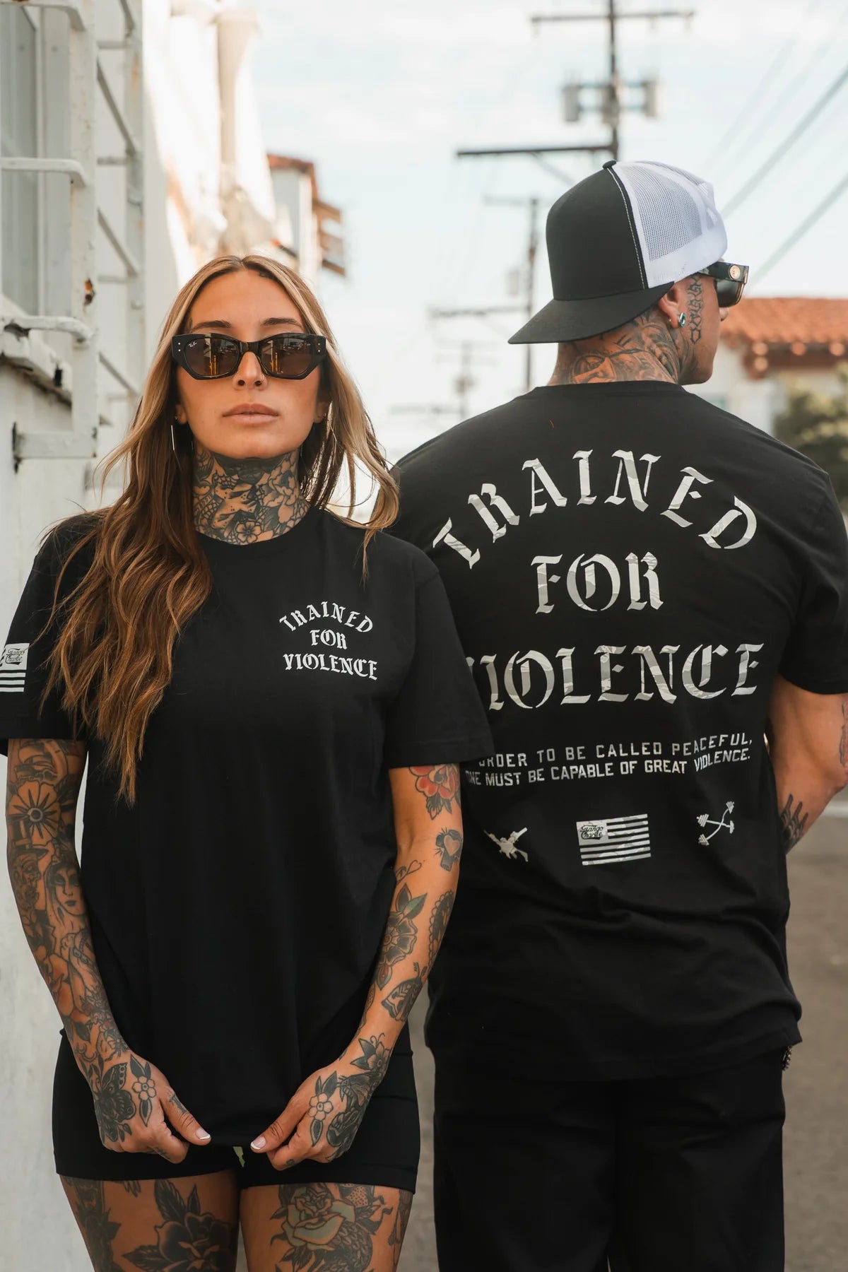 Two tattooed people wear the Trained for Violence - Tee by Tango Charlie Apparel—one faces forward in sunglasses, the other shows off the back design as they stand outside on a sunny day.