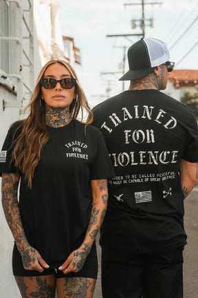 Two tattooed people wear the Trained for Violence - Tee by Tango Charlie Apparel—one faces forward in sunglasses, the other shows off the back design as they stand outside on a sunny day.