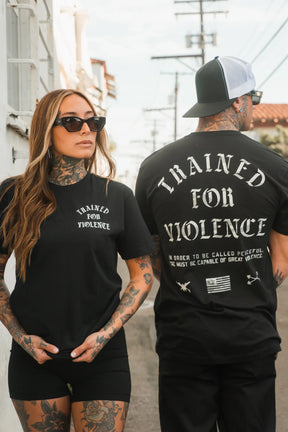 A woman and man wear black Trained for Violence - Tee shirts by Tango Charlie Apparel outdoors, embodying psychological conditioning. The man's shirt features a longer quote on the back as they stand facing different directions.