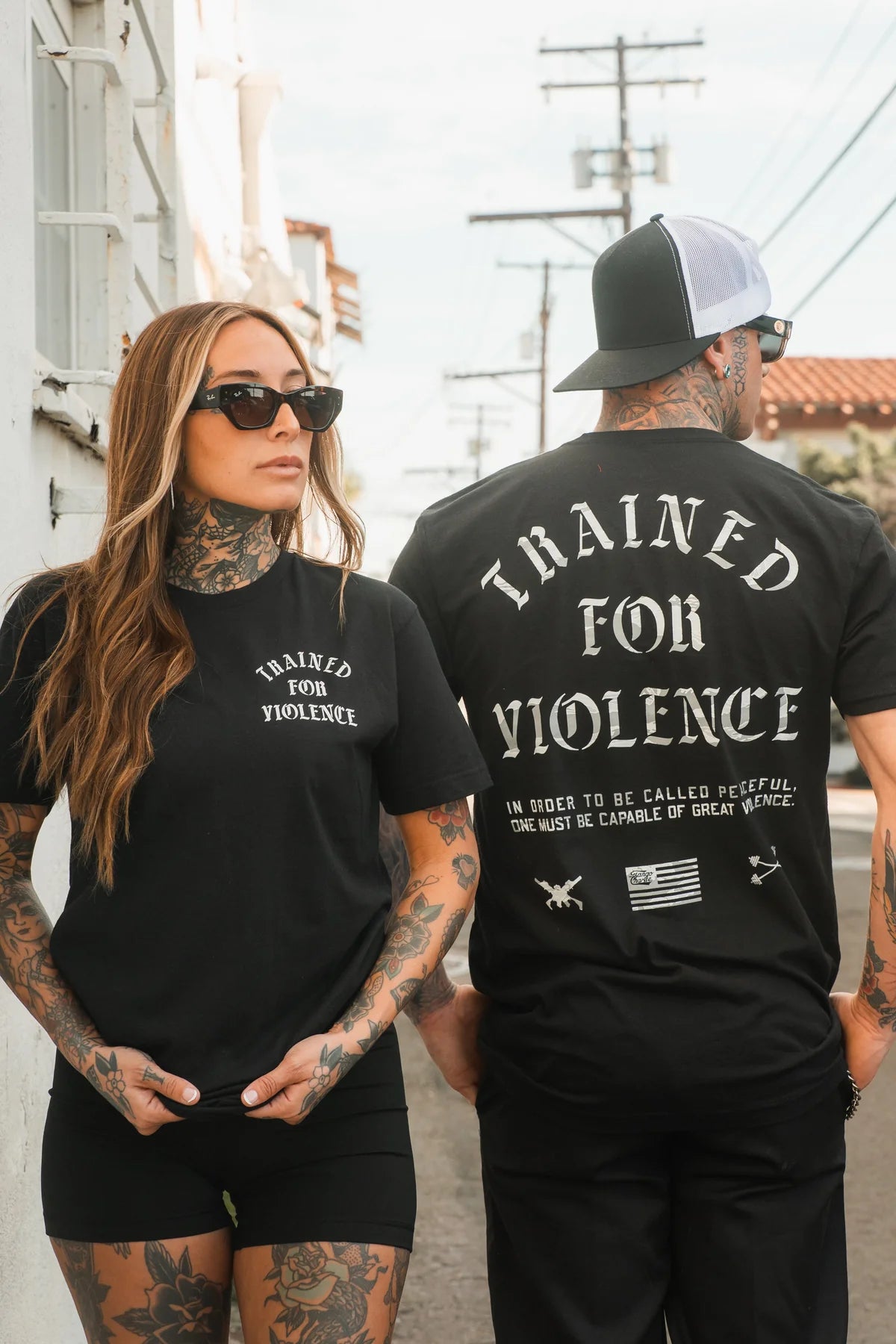 Two tattooed individuals model the "Trained for Violence - Tee" by Tango Charlie Apparel outdoors—the woman faces forward in sunglasses, while the man shows off the tee's bold back design, embodying strength and style.