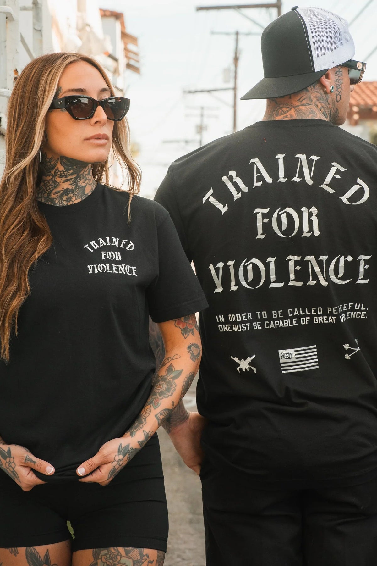 A tattooed man and woman wear Tango Charlie Apparel’s Trained for Violence Tee. Hers displays the bold slogan, while his shirt’s back states that true peace demands both strength and technical proficiency.
