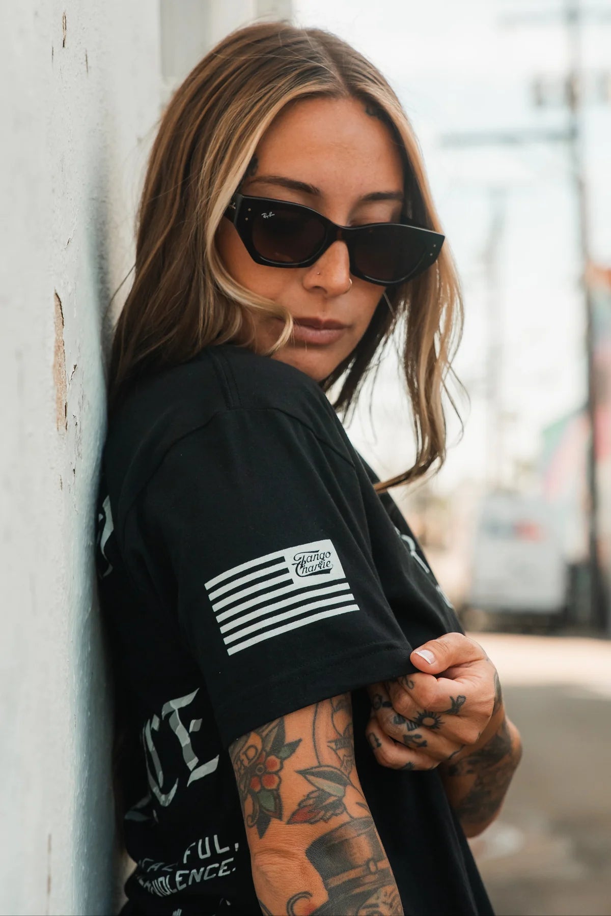 A woman with long, light brown hair and sunglasses leans against a wall, revealing her tattooed arm and the striped patch on her Trained for Violence - Tee by Tango Charlie Apparel, exuding technical skill in an urban backdrop.