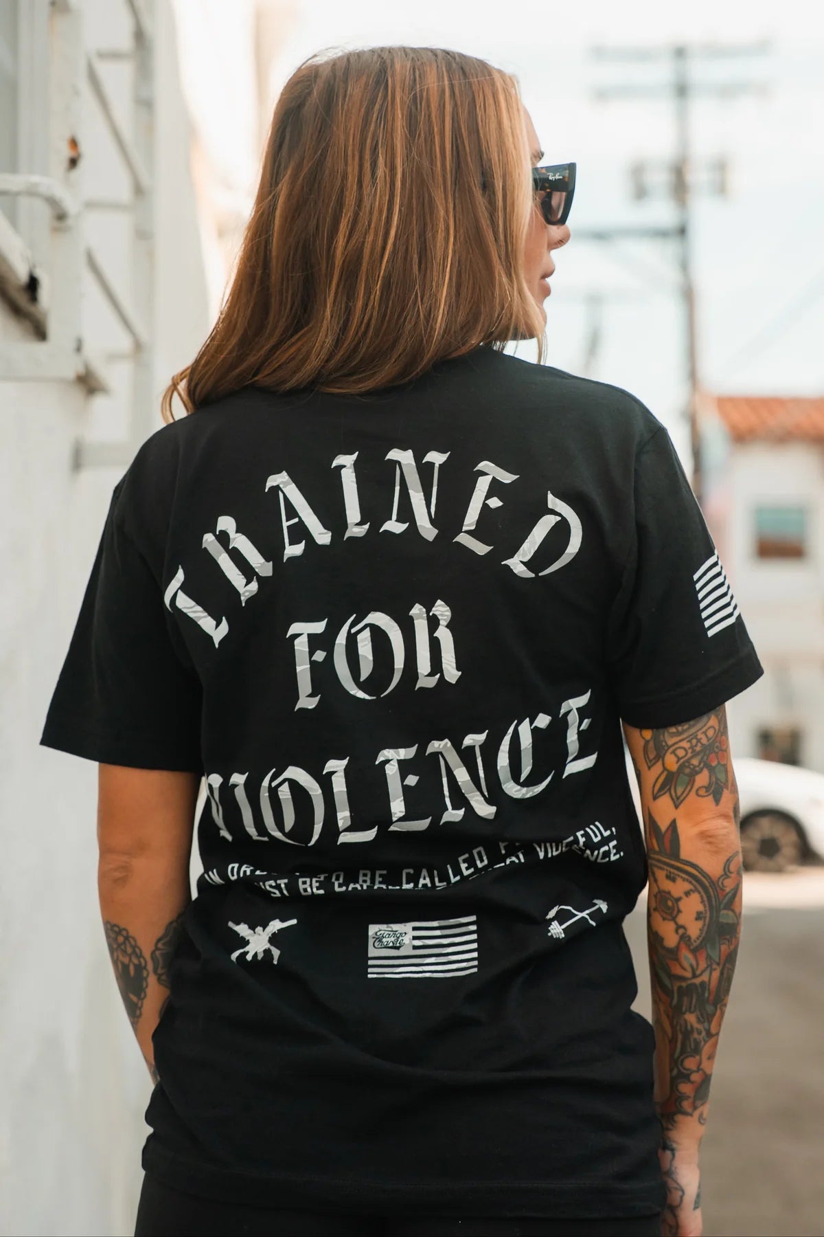 A tattooed woman in sunglasses wears a Tango Charlie Apparel "Trained for Violence - Tee" as she stands outdoors in daylight, framed by buildings and power lines.