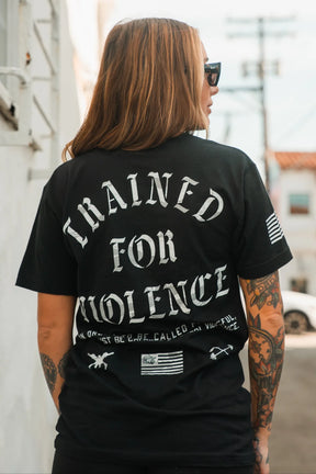 A tattooed woman in sunglasses wears a Tango Charlie Apparel "Trained for Violence - Tee" as she stands outdoors in daylight, framed by buildings and power lines.