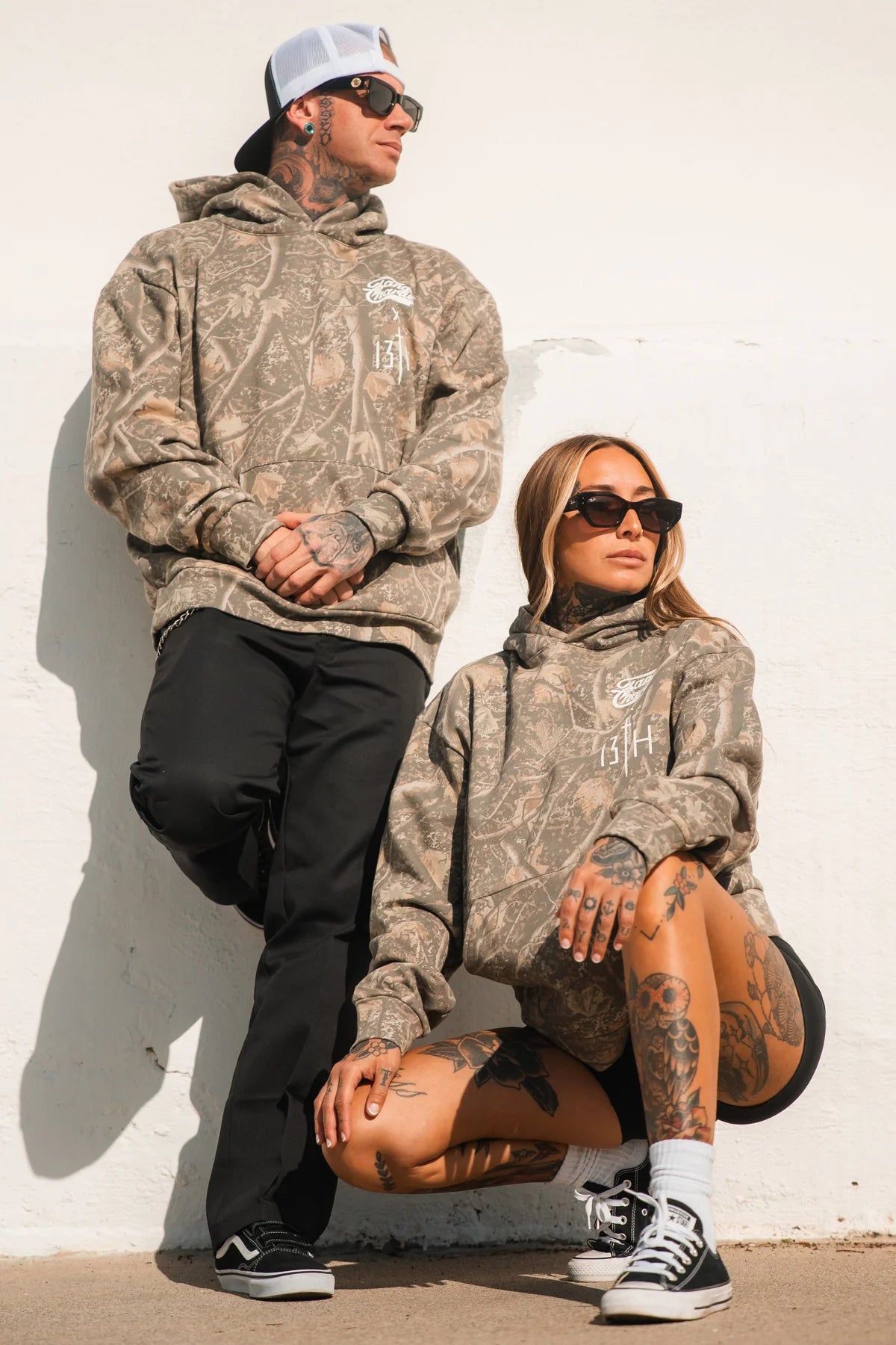 Two people in Lift Heavy. Pray Hard. - Deadwood Camo Hoodies by Tango Charlie Apparel pose confidently—one standing, one kneeling—in black sunglasses and sneakers against a white wall.