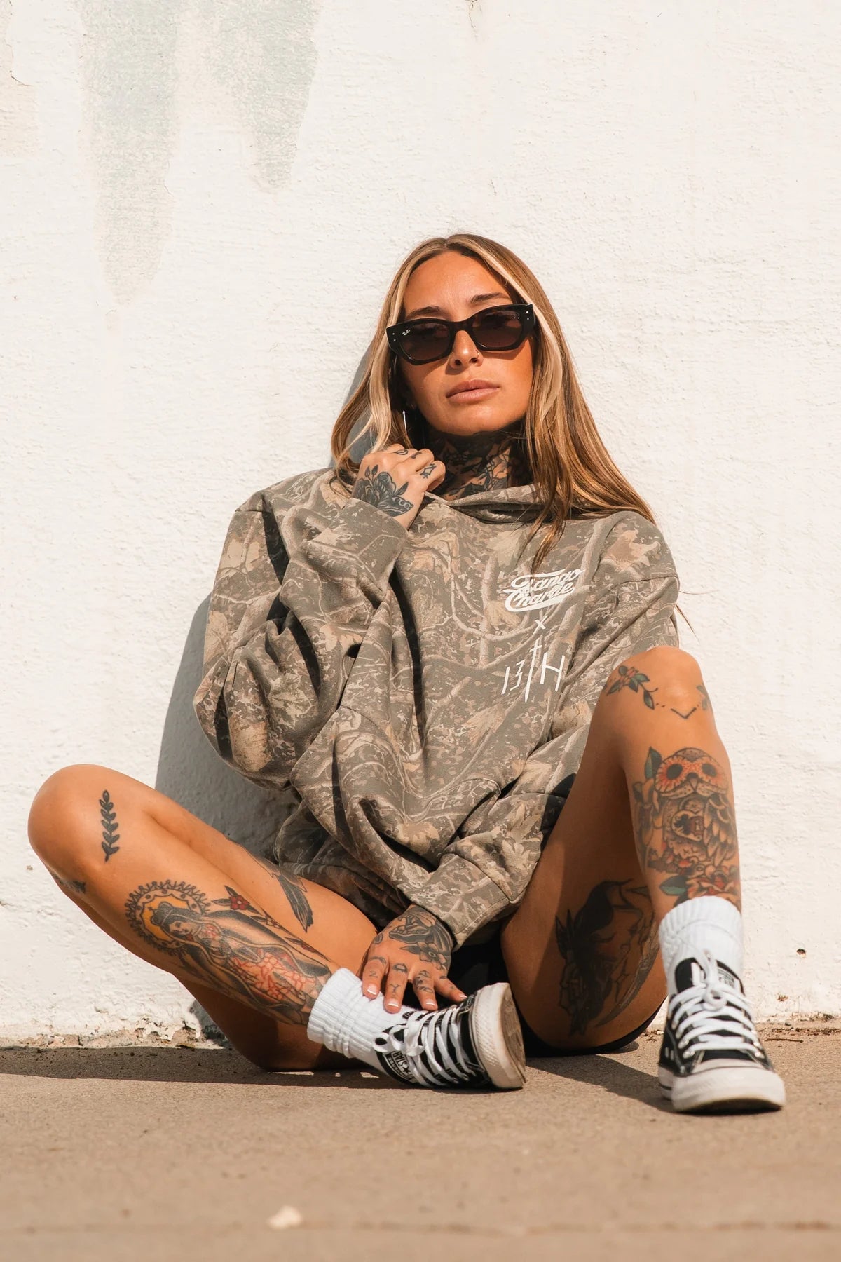A woman in the Lift Heavy. Pray Hard. - Deadwood Camo Hoodie by Tango Charlie Apparel, sunglasses, and black sneakers sits against a white wall, exuding confident, spiritual vibes.