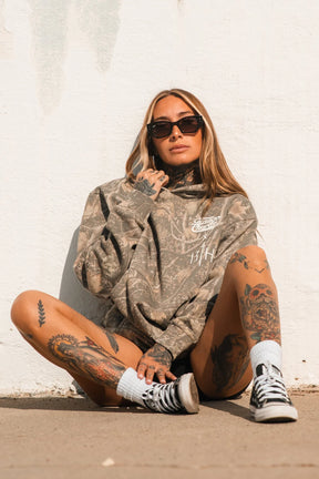 A woman in the Lift Heavy. Pray Hard. - Deadwood Camo Hoodie by Tango Charlie Apparel, sunglasses, and black sneakers sits against a white wall, exuding confident, spiritual vibes.
