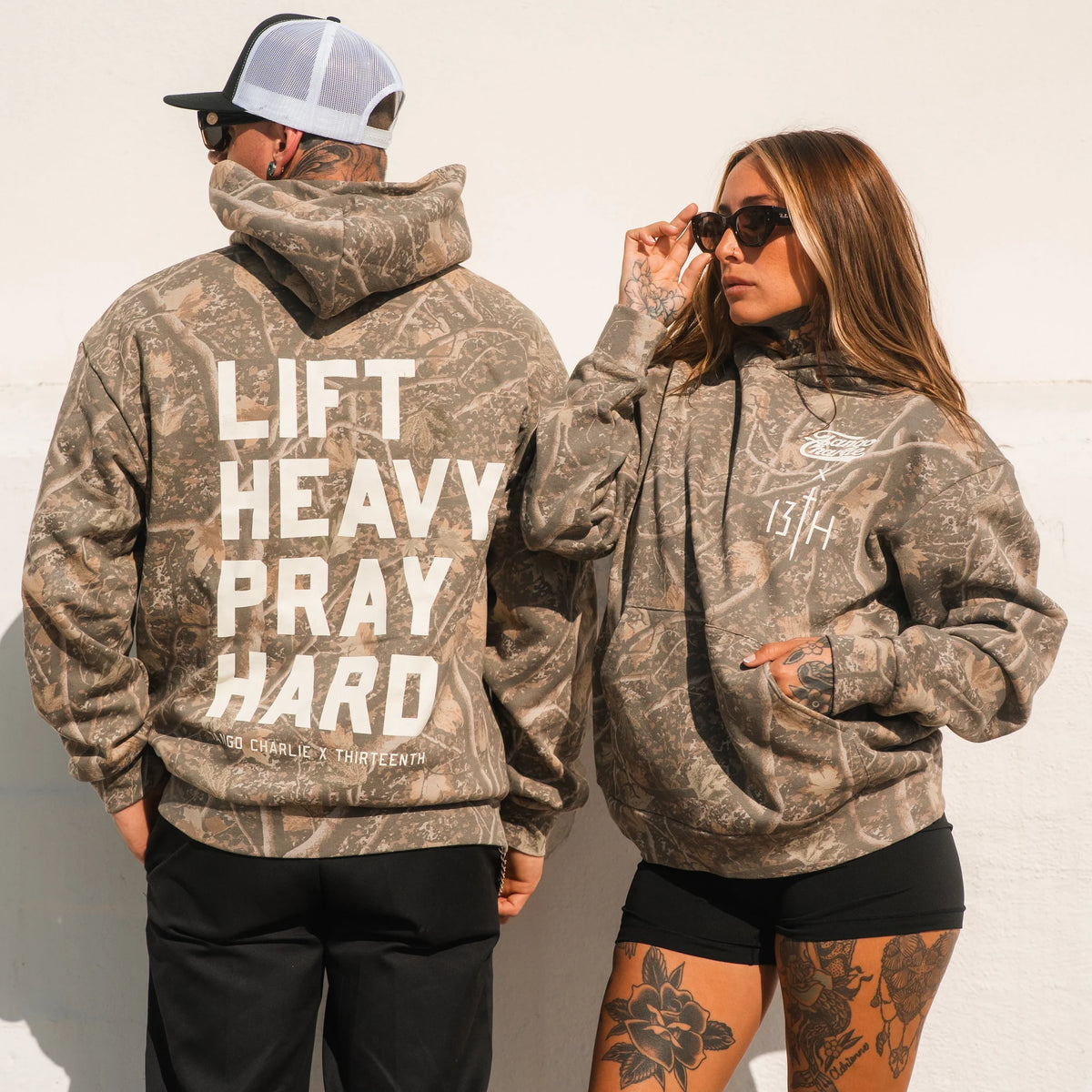 Two people in Lift Heavy. Pray Hard. - Deadwood Camo Hoodie by Tango Charlie Apparel wear sunglasses and pose against a white wall, embodying bold, spiritual strength.