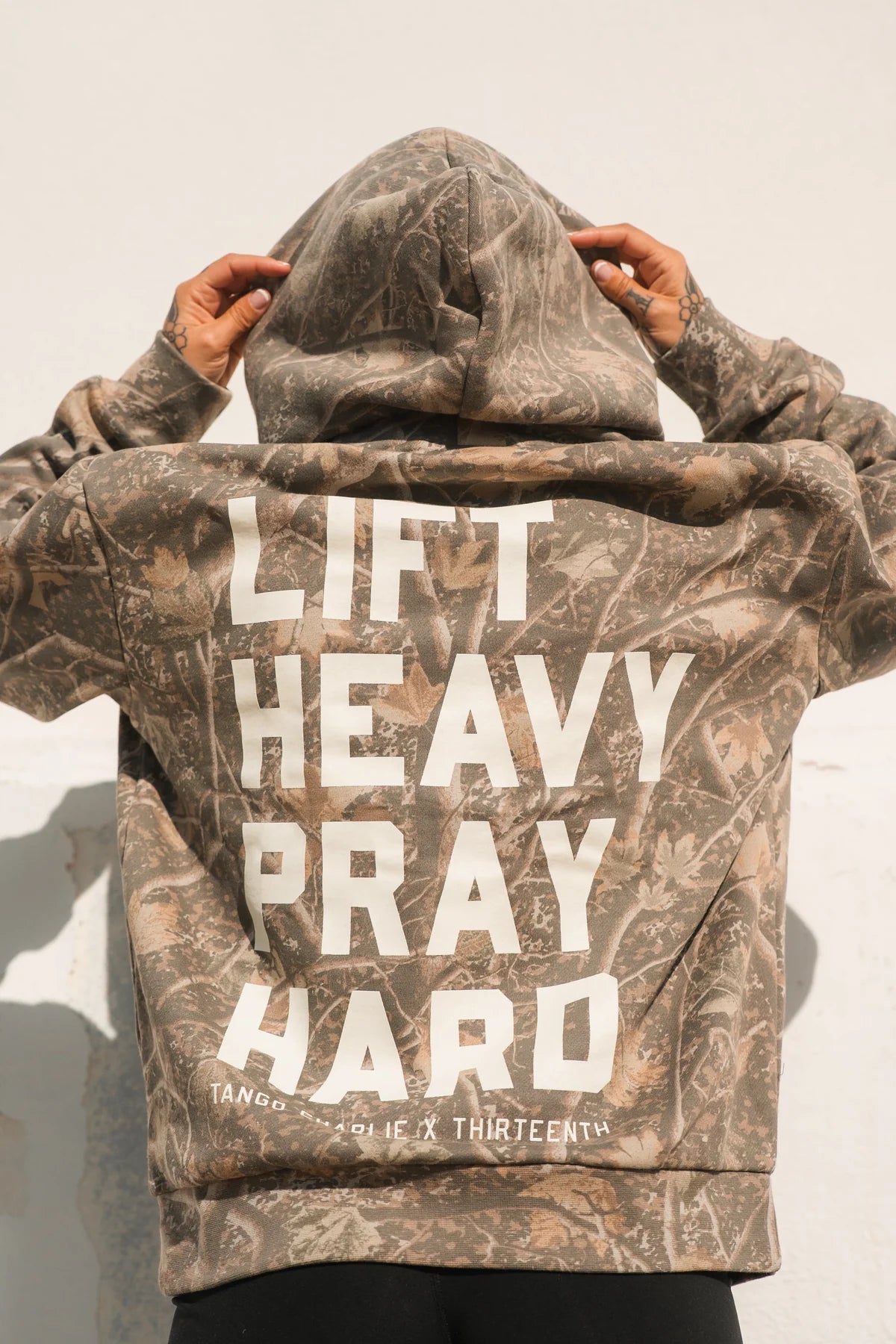 A person faces away, hands on hood, wearing the Lift Heavy. Pray Hard. - Deadwood Camo Hoodie by Tango Charlie Apparel, embodying spiritual strength against a light background.
