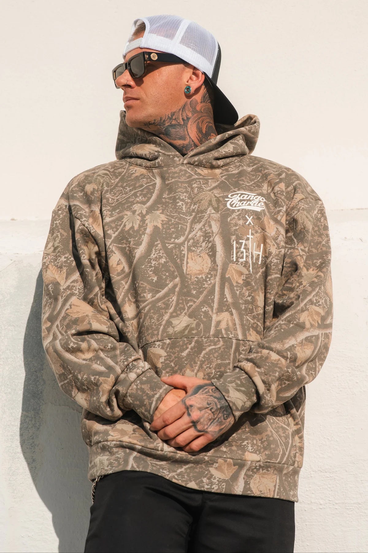 A man in the Lift Heavy. Pray Hard. - Deadwood Camo Hoodie by Tango Charlie Apparel, black pants, sunglasses, and a white mesh cap stands against a light wall, looking to the side.