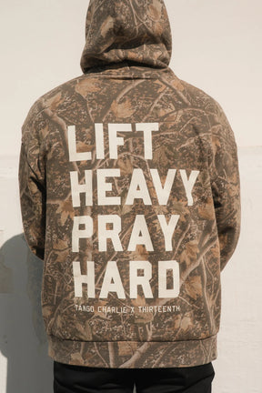 Person wearing the Lift Heavy. Pray Hard. - Deadwood Camo Hoodie by Tango Charlie Apparel, embodying resilience and spiritual strength in style.