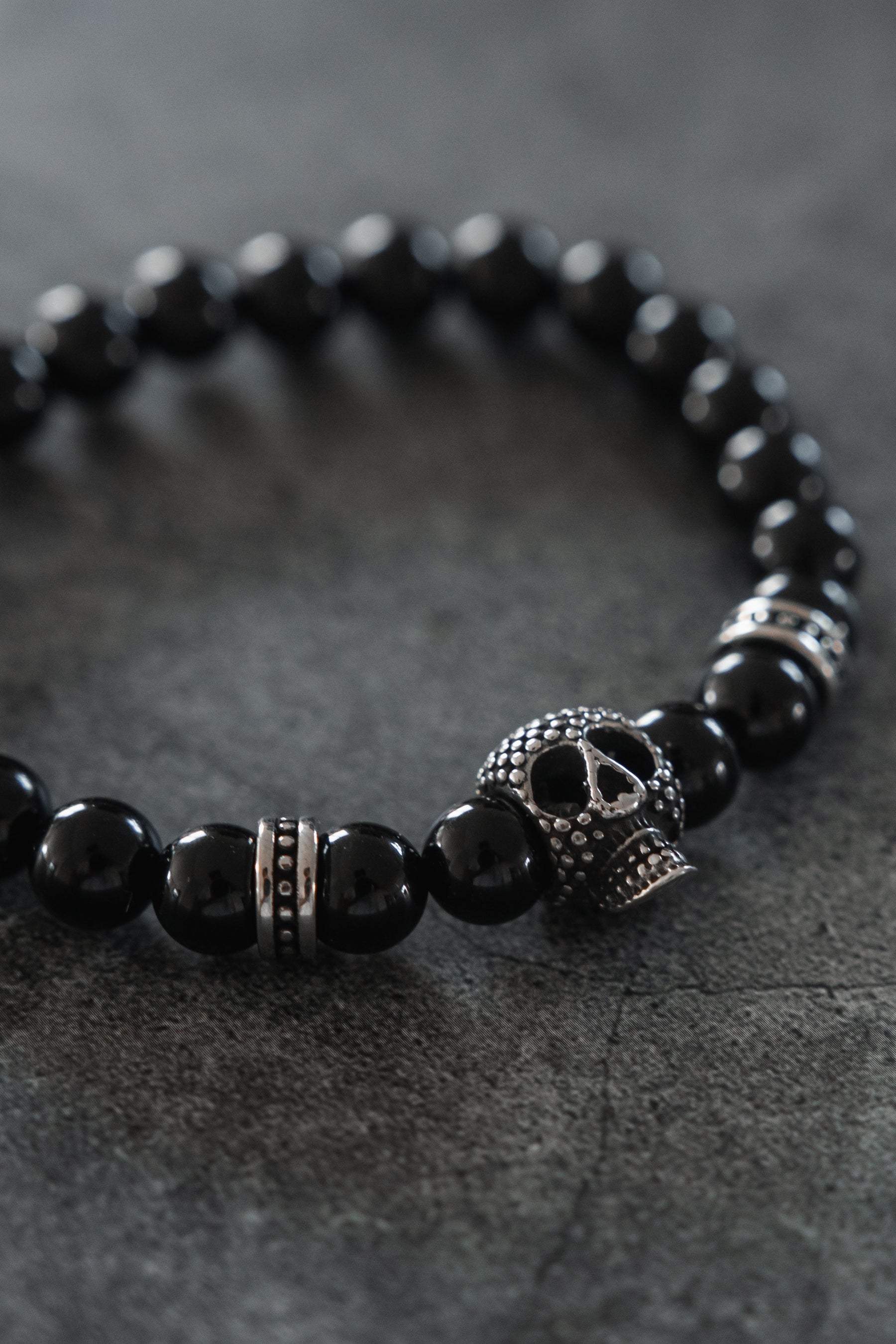 Introducing the Relentless Betrayal Day of the Dead Black Bead Bracelet — a bold accessory with round black beads and metallic accents, highlighted by an intricately crafted metal skull bead that channels the iconic Day of the Dead style, beautifully displayed on a dark textured surface.