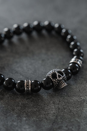 Introducing the Relentless Betrayal Day of the Dead Black Bead Bracelet — a bold accessory with round black beads and metallic accents, highlighted by an intricately crafted metal skull bead that channels the iconic Day of the Dead style, beautifully displayed on a dark textured surface.
