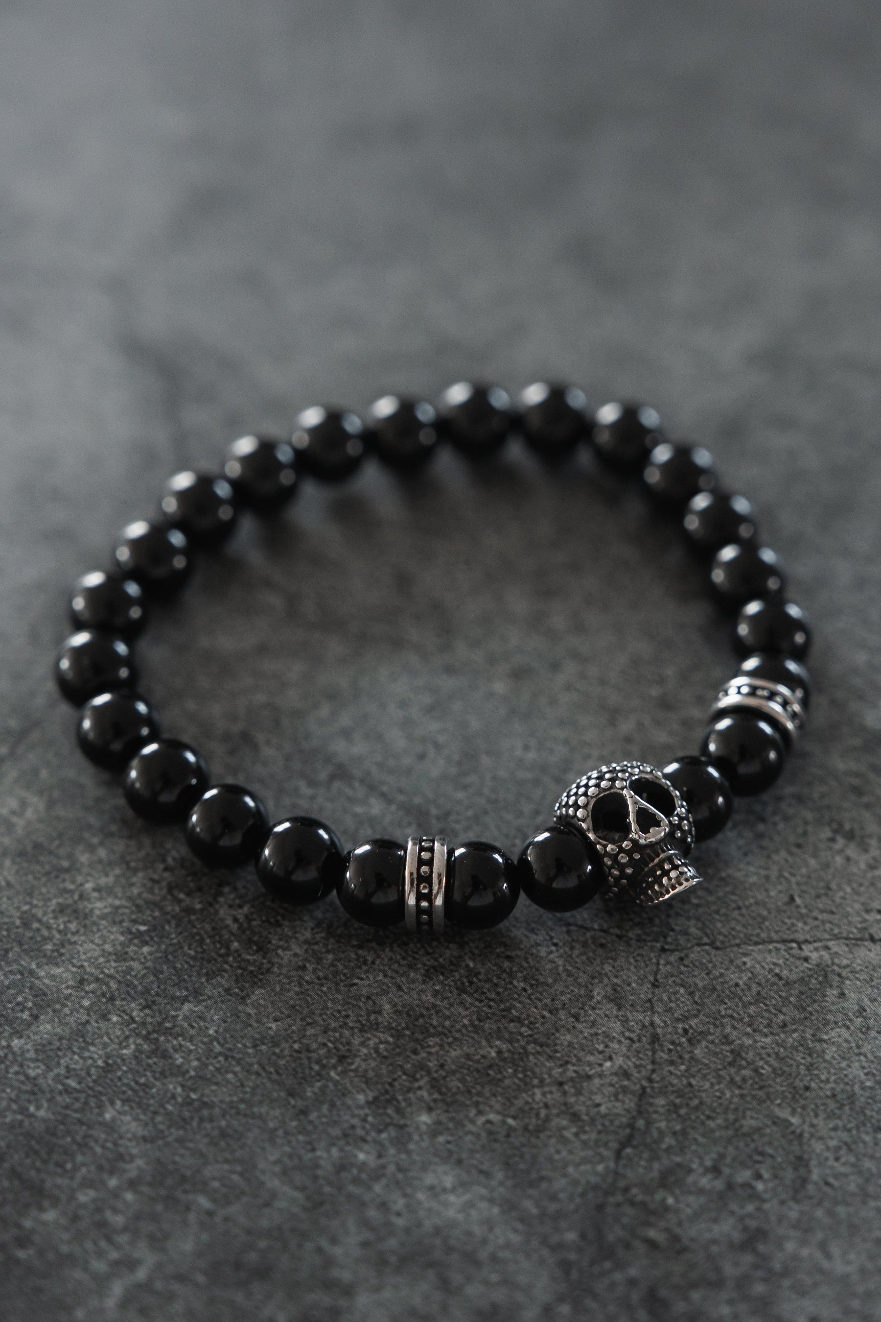 The Relentless Betrayal Day of the Dead Black Bead Bracelet features semi-precious gemstones, silver-toned accents, and a decorative skull charm inspired by Day of the Dead designs, displayed on a dark, textured surface.