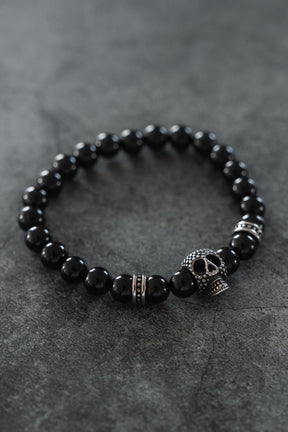 The Relentless Betrayal Day of the Dead Black Bead Bracelet features semi-precious gemstones, silver-toned accents, and a decorative skull charm inspired by Day of the Dead designs, displayed on a dark, textured surface.