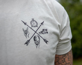 Close-up of the Doers Drifters Lifters Punks Unisex Tee by Tango Charlie Apparel, featuring two crossed arrows with a clock, sneaker, rock hand sign, and fist graphic. Tattooed arm partly visible—rep your crew in style.