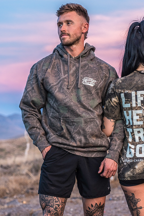 A man stands outdoors at dusk, embodying the spirit of Christian Fitness in the Tango Charlie Apparel “Lift Heavy. Trust God.” Premium Darkwood Camo Hoodie and black shorts; beside him, another individual in a graphic shirt is partially visible.
