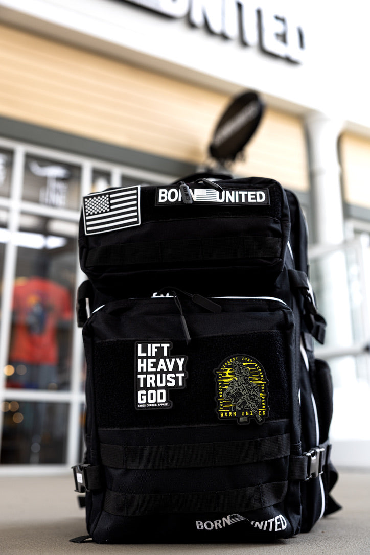 The Born United Private Label Apex Premium Tac-Pac - 45L, a black tactical travel backpack with patches like an American flag, BORN UNITED, LIFT HEAVY TRUST GOD, and a yellow design, rests outside a building with large windows and a partial UNITED sign.
